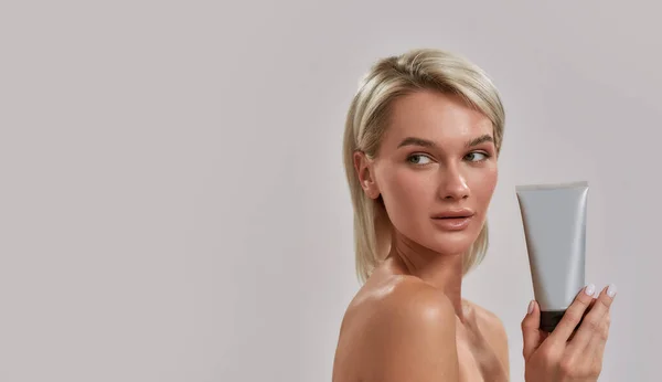 Portrait of young woman with perfect fresh skin looking aside, holding cosmetic skincare, bodycare product while posing isolated over grey background — Stock Photo, Image