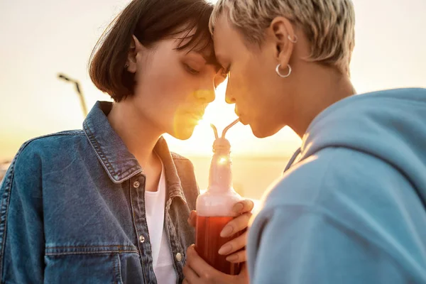 Cropped shot of happy young lesbian couple drinking from one glass bottle with the straw, Two girls enjoying cold beverage on a summer day outdoors — Stock Photo, Image