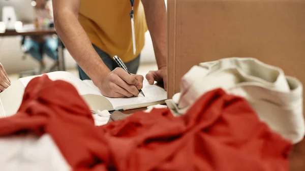 Cropped shot of young male volunteer making notes while checking clothes in donation box for needy people, Selective focus on hand — Stock Photo, Image