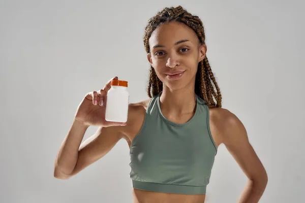 Primo piano della giovane donna felice corsa mista fitness in possesso di bottiglia con integratori nutrizionali sportivi e sorridente alla fotocamera mentre in piedi isolato su sfondo grigio — Foto Stock