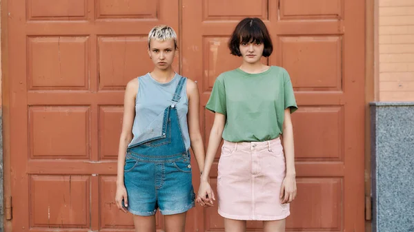 Pareja femenina joven, dos chicas mirando a la cámara, cogidas de la mano mientras están de pie frente a la puerta al aire libre. LGBT, Concepto de libertad sexual — Foto de Stock