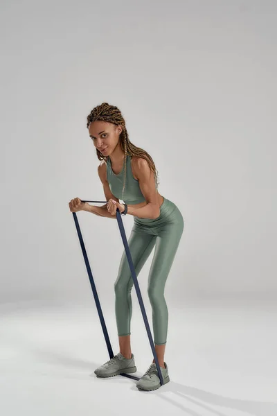 Trainingsbenen. Verticale opname van een jonge atletische gemengde ras vrouw in sportkleding geïsoleerd over grijze achtergrond en oefenen met elastische band of fitness gom — Stockfoto