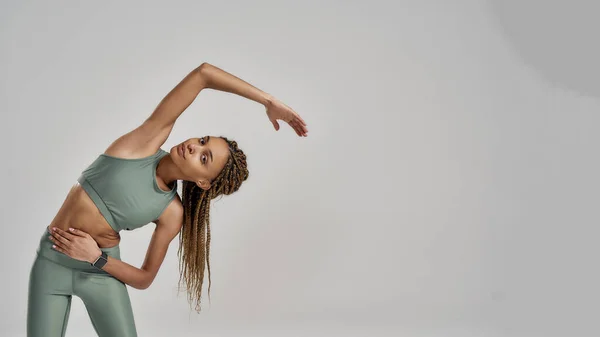 Opwarmen. Jonge gemengde ras fitness vrouw in sportkleding oefening terwijl geïsoleerd over grijze achtergrond, kopieer ruimte voor tekst — Stockfoto