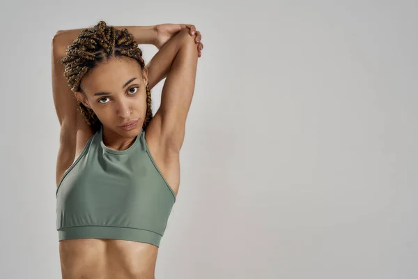 Buena rutina de calentamiento. Joven mujer de raza mixta en ropa deportiva estirando los brazos antes de entrenar fitness y mirando a la cámara mientras está aislada sobre un fondo gris — Foto de Stock