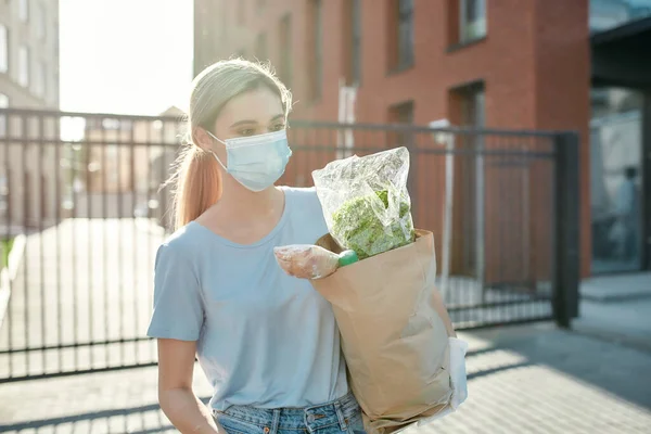 Mujer joven con máscara médica y guantes protectores caminando con la bolsa de comestibles a su coche después de hacer compras en un supermercado —  Fotos de Stock