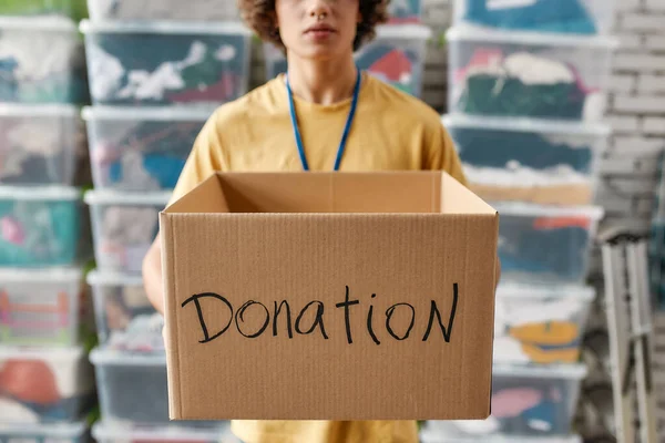 Beskuren bild av en kille som håller i donationslådan, poserar framför lådor fulla med kläder, Ung frivillig man som arbetar för en välgörenhet, Fokus på pappkartong — Stockfoto