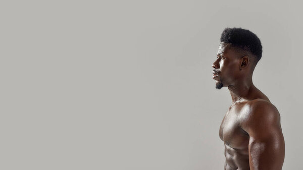 Young muscular african american man with naked torso looking away while posing shirtless isolated over grey background. Sports, workout, bodybuilding concept. Side view. Web Banner