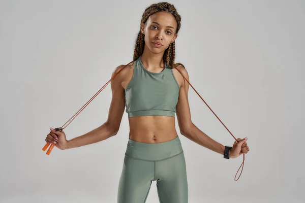 Portrait of young athletic mixed race woman in sportswear holding skipping rope and looking at camera while standing against grey background in studio — Stock Photo, Image