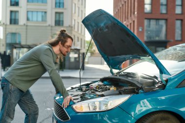 Odaklanmış görünen genç adam, şehir caddesinde dururken kırık arabasını açık kaputla inceliyor.