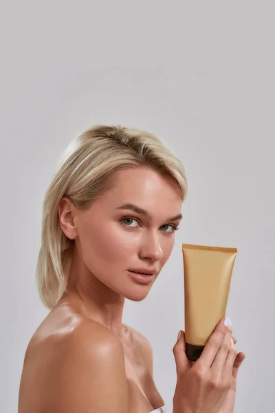 Porträt einer jungen Frau mit perfekt glühender Haut, die in die Kamera blickt, Sonnencreme, Bronzer, Pflegeprodukt in der Hand hält und isoliert vor grauem Hintergrund posiert — Stockfoto