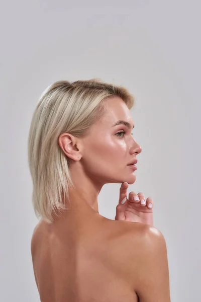 Porträtt av vackra unga kvinnliga blond med perfekt fräsch hud tittar åt sidan, vidrör hakan samtidigt poserar isolerad över grå bakgrund — Stockfoto