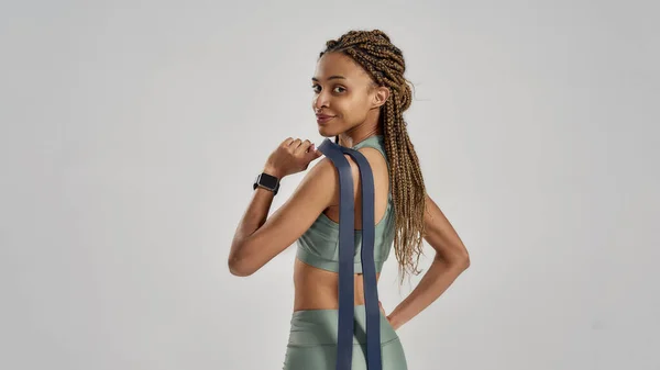 Ruhe nach dem Training. Schöne junge Mischlingsfrau mit Gummiband oder Fitnessgummi und lächelnd in die Kamera, während sie isoliert vor grauem Hintergrund posiert, Rückansicht — Stockfoto