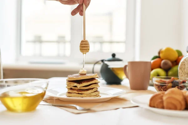 Hand holding dipper, adding honey on top of stack of sweet tasty pancakes, Teapot, croissants and various fruits served for breakfast on the table — Stock Photo, Image