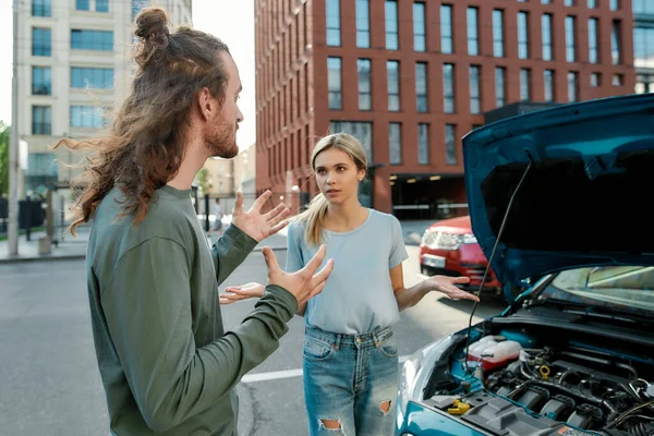 Portret van een jonge blanke man en vrouw ruziënd, bij de kapotte auto met open motorkap op straat — Stockfoto