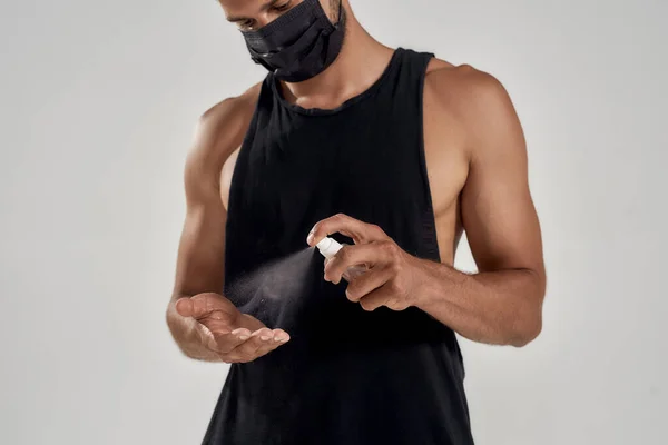 Safety. Young muscular man in medical mask using hand sanitizer while standing isolated over grey background — Stock Photo, Image