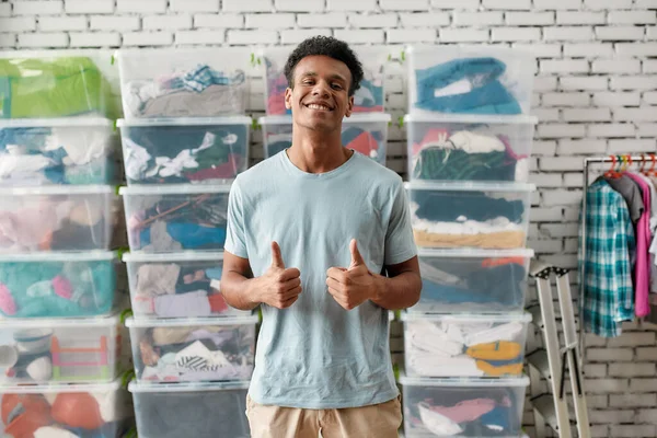 Vrolijke man lacht naar de camera, toont duimen omhoog, poseert voor rek en dozen vol kleren, Jonge mannelijke vrijwilliger werkt voor een goed doel, doneert kleding aan behoeftige mensen — Stockfoto