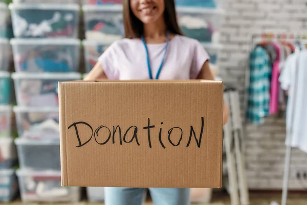 Foto recortada de una mujer sonriente sosteniendo la caja de donación, posando frente a un estante y cajas llenas de ropa, Joven voluntaria trabajando para una organización benéfica, Focus on cardboard box — Foto de Stock