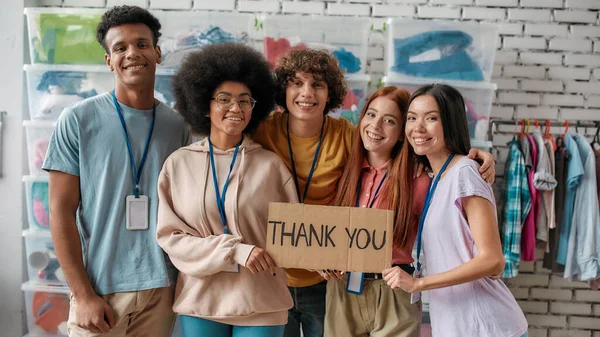 Grupo de jóvenes voluntarios alegres y diversos sonriendo a la cámara, sosteniendo la tarjeta con letras de agradecimiento mientras posan en la oficina de la organización caritativa — Foto de Stock