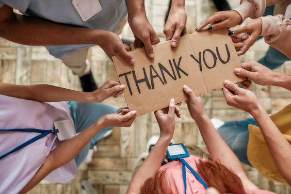 Top view of diverse young volunteers holding card with Thank you lettering while standing in charitable organization office