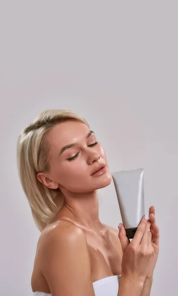 Retrato de mujer joven con piel fresca perfecta que sostiene el cuidado de la piel cosmética, producto de cuidado corporal mientras posa aislado sobre fondo gris — Foto de Stock