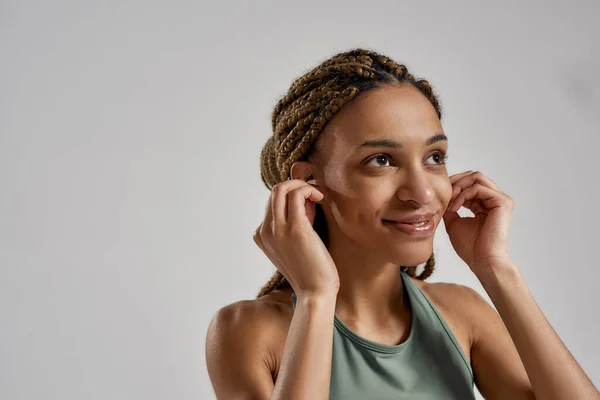 Musica per allenamento. Ritratto di giovane donna felice mista corsa fitness indossando auricolari wireless e sorridente mentre in piedi isolato su sfondo grigio — Foto Stock