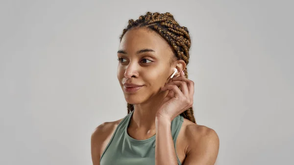 Ascolto musica. Ritratto di giovane attraente donna di fitness mista che indossa auricolari wireless e sorride in piedi isolata su sfondo grigio — Foto Stock
