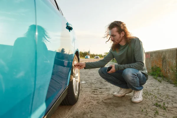 Young caucasian man checking air pressure of car tire on local road side while traveling, Male driver having troubles with his auto, checking wheel after car breakdown — Stock Photo, Image