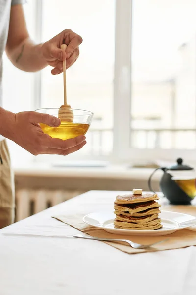 Close-up van de handen van de man houden dipper in een kom met honing tijdens het bereiden van het ontbijt, Een stapel zoete smakelijke pannenkoeken en theepot geserveerd op de tafel — Stockfoto