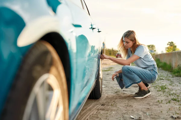 Aantrekkelijke jonge vrouw controleren luchtdruk van autoband op de lokale weg tijdens het reizen, Meisje heeft problemen met haar auto, het controleren van wiel na autopech — Stockfoto