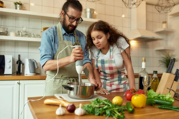 Італієць, шеф-кухар за допомогою ручного блендера під час приготування їжі. Молода жінка, дівчина в фартусі, дивиться на процес, допомагає на кухні. — стокове фото