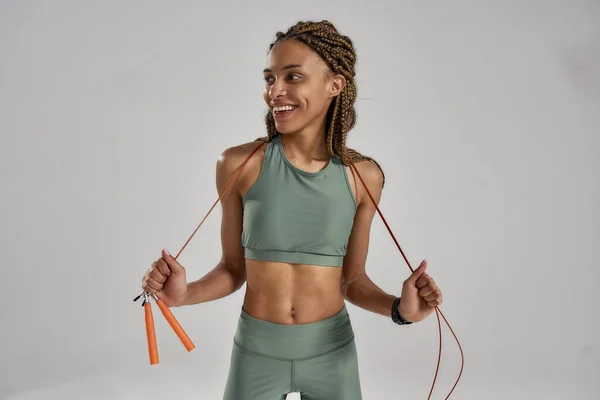 Joven en forma y saludable mujer de raza mixta en ropa deportiva sosteniendo saltar la cuerda, mirando a un lado y sonriendo mientras está de pie sobre fondo gris en el estudio — Foto de Stock