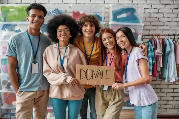 Grupa radosnych, różnorodnych młodych wolontariuszy uśmiechnięta do kamery, trzymająca kartkę z literami Donate stojąc w biurze organizacji charytatywnej — Zdjęcie stockowe