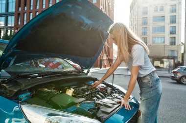 Şehir caddesinde duran genç bir kadın bozuk arabasını açık kaputla inceliyor.