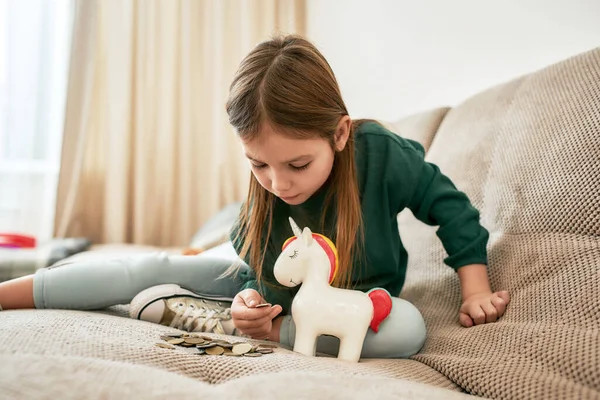 Una niña pequeña en un jersey verde está sentada sola en un sofá examinando monedas que tienen su monedero unicornio alrededor de sdgsdfg —  Fotos de Stock