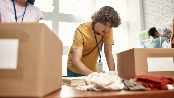 Young volunteers sorting, packing clothes in cardboard boxes, Diverse team working on donation project indoors, Focus on a guy — Stock Photo, Image