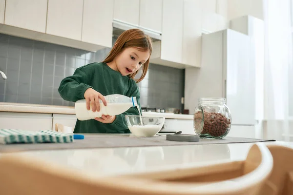 Ein kleines nettes Mädchen hält eine Flasche Milch und gießt sie in eine Schüssel, während sie in einer Küche steht — Stockfoto