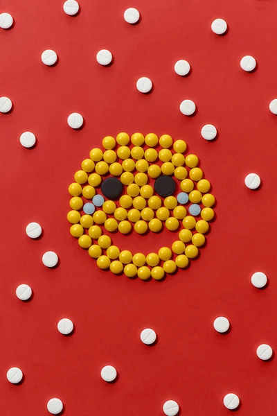 Visão superior de diferentes comprimidos redondos coloridos que se encontram na forma de emoji, rosto isolado sobre fundo vermelho. Conceito de cuidados de saúde e tratamento. — Fotografia de Stock