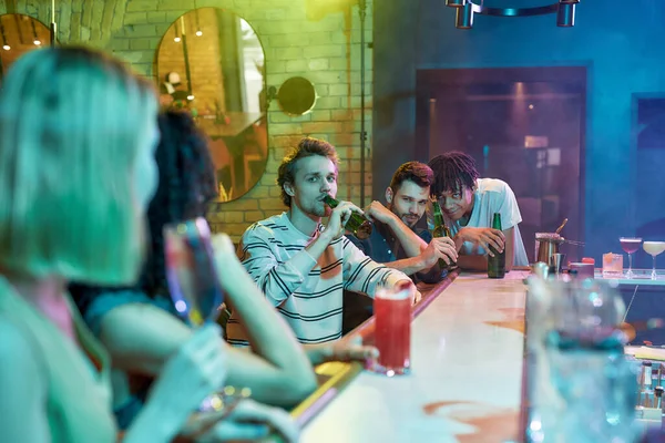 Tre fyre drikker øl, flirter, kigger på kvinder, der sidder ved bardisken. Venner bruger tid på natklub, restaurant - Stock-foto