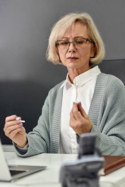 Yaşlanmış kadın portresi, kıdemli stajyer, kablosuz kulaklıklara bakmak, dizüstü bilgisayar kullanmak, masa başında oturmak, modern ofiste çalışmak.