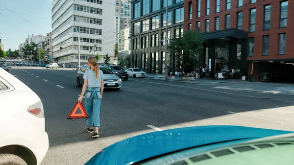 Full length shot of young woman walking on the city street, putting red triangle emergency warning sign on the road while having problems with her car — Stock Photo, Image