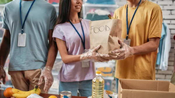 Foto recortada de voluntarios masculinos y femeninos sosteniendo bolsa de papel con comida mientras empacan la donación para personas necesitadas, equipo joven que trabaja en la fundación benéfica — Foto de Stock