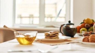 Bir yığın tatlı krep, bir kase içinde bal, çaydanlık, kruvasan ve masada kahvaltıda servis edilen çeşitli meyveler.