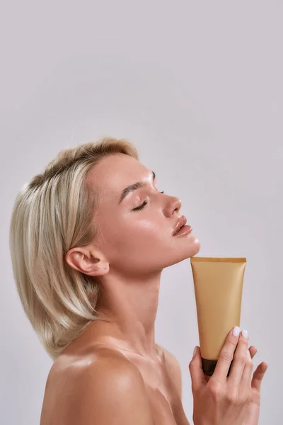 Retrato de mulher jovem com pele brilhante perfeita posando com os olhos fechados, segurando protetor solar, bronzeador, produto de cuidados com a pele enquanto está isolada sobre fundo cinza — Fotografia de Stock