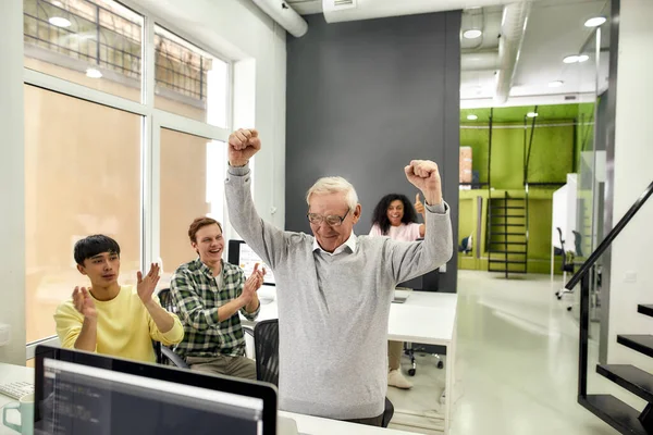 Åldrad man, seniorpraktikant ser glad ut, höjer armarna efter att ha avslutat sin första uppgift på jobbet, Vänliga arbetare applåderar, hejar på ny anställd på kontoret — Stockfoto