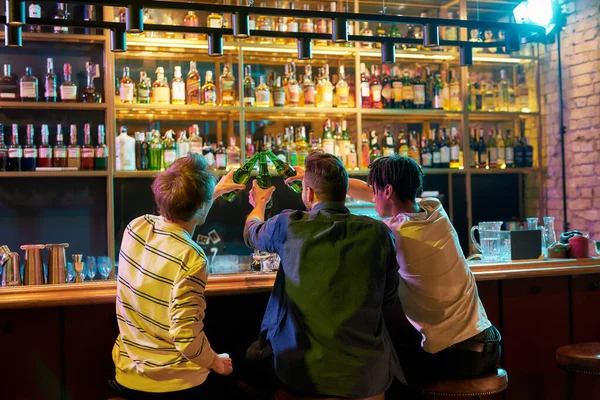 Buen momento con los amigos. Tres hombres tintinean botellas de cerveza mientras están sentados en el mostrador del bar. Amigos pasando tiempo en el bar de deportes — Foto de Stock