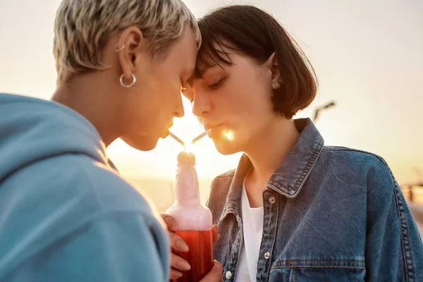 Cropped shot of young lesbian couple having romantic moment, drinking from one glass bottle with the straw, Two women enjoying cold beverage on a summer day outdoors — Stock Photo, Image