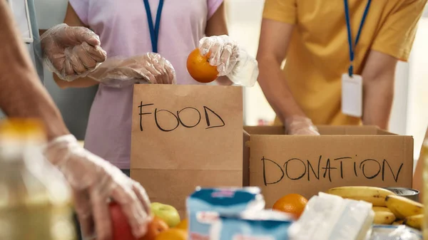 Närbild av händerna på frivilliga i handskar samla in, sortera mat för behövande människor i papperspåsar, Team som arbetar tillsammans med donationsprojekt — Stockfoto