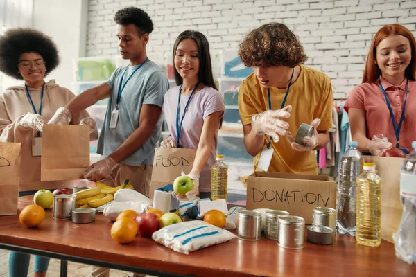 Jonge mannelijke en vrouwelijke vrijwilligers die voedsel en dranken doneren voor daklozen in papieren zakken, Kleine groep gelukkige mensen die werken in liefdadigheidsstichting — Stockfoto