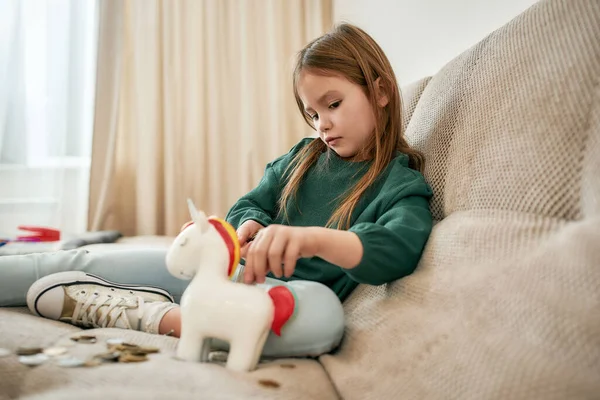 Una niña pequeña en un suéter verde está llenando su monedero de unicornio con monedas una por una en un sofá beige —  Fotos de Stock