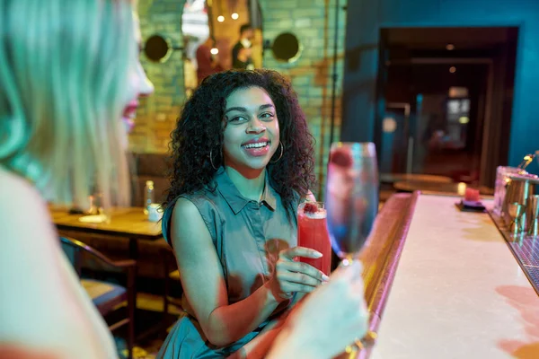 Dos mujeres divirtiéndose, brindando, bebiendo cócteles mientras están sentadas en el mostrador del bar. Amigos pasando tiempo en el club nocturno, restaurante — Foto de Stock